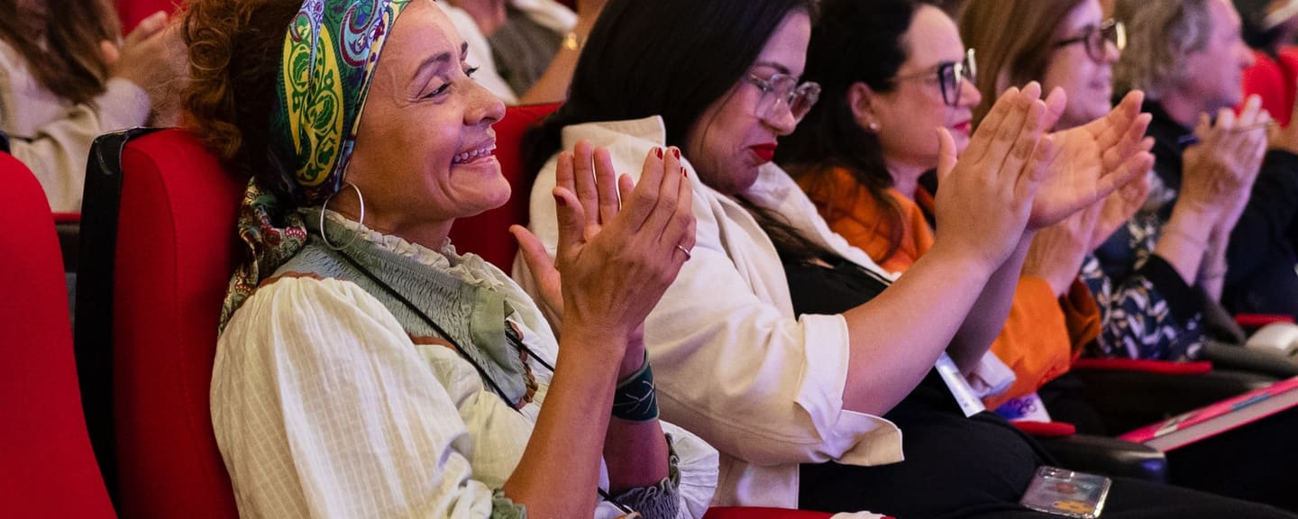 Mulher de lenço estampado sorri e aplaude na plateia do II Fórum, com outras participantes aplaudindo ao lado