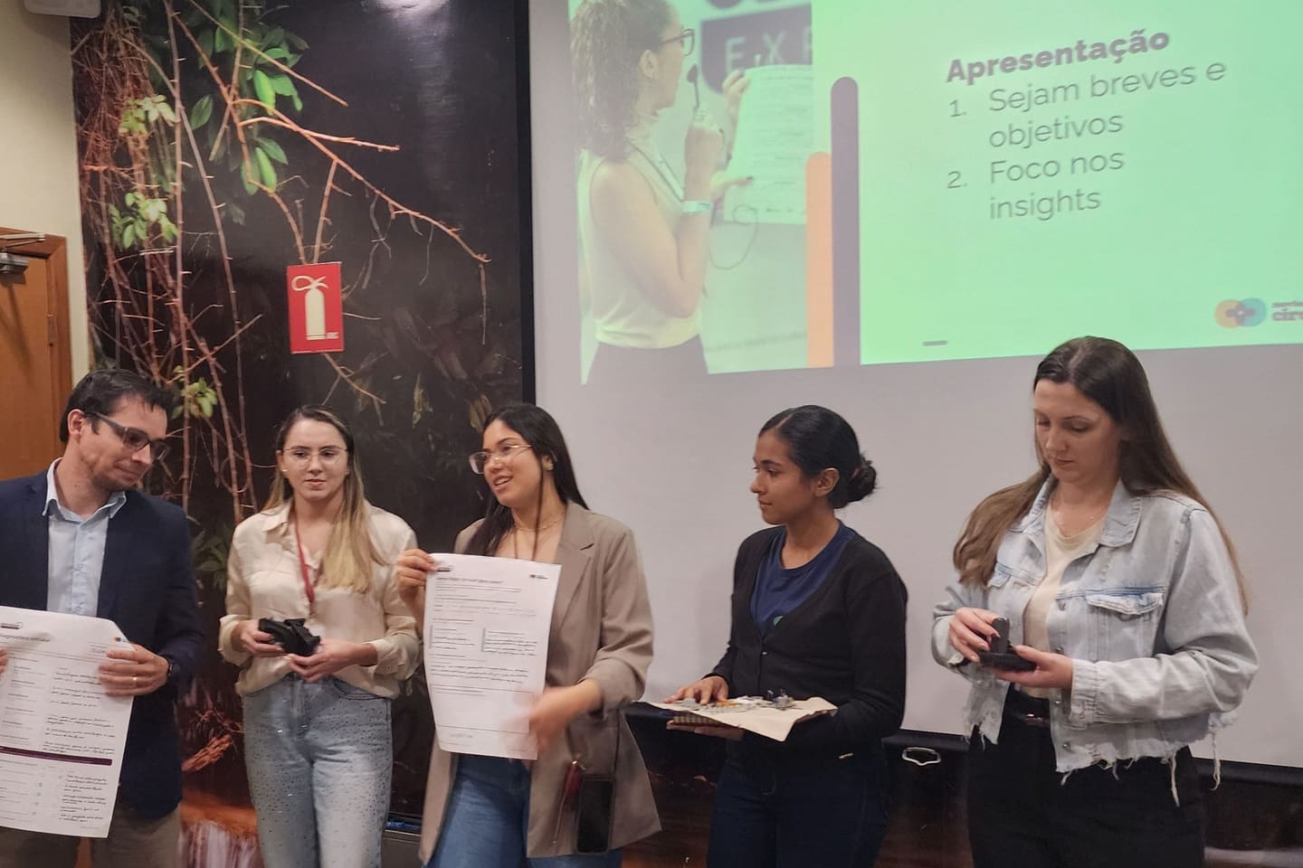 Cinco participantes em pé, lado a lado, segurando os canvas preenchidos e as peças que desmontaram, apresentando para a sala diante do slide de apresentação