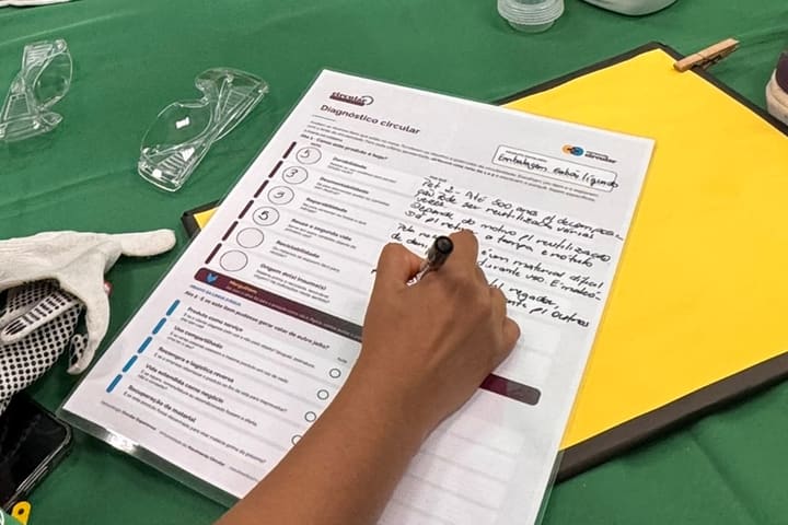 Mão preenchendo à caneta a ficha do Diagnóstico circular, com as notas nos seis critérios e os porquês escritos, ao lado da luva e dos óculos de proteção