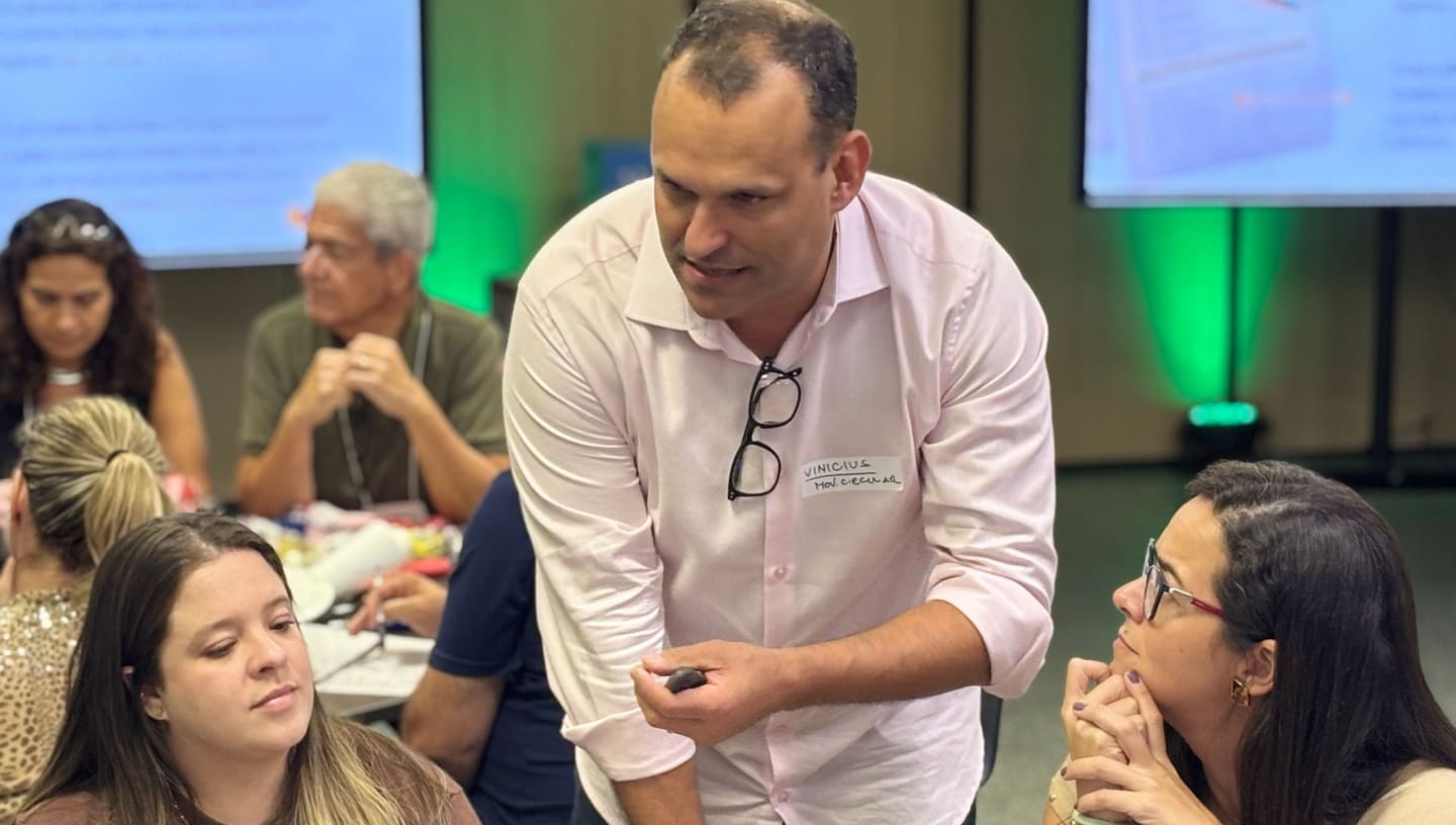 O facilitador do Movimento Circular, inclinado sobre a mesa com uma peça desmontada na mão, conversa com uma participante que escuta com a mão no queixo, durante a oficina